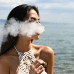 a woman with black hair vaping and exhaling smoke in the air near the sea